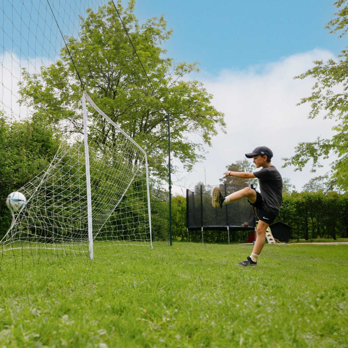 young boy practicing soccer kicking ball into 3 in 1 Soccer Goal Trainer in backyard on grass field