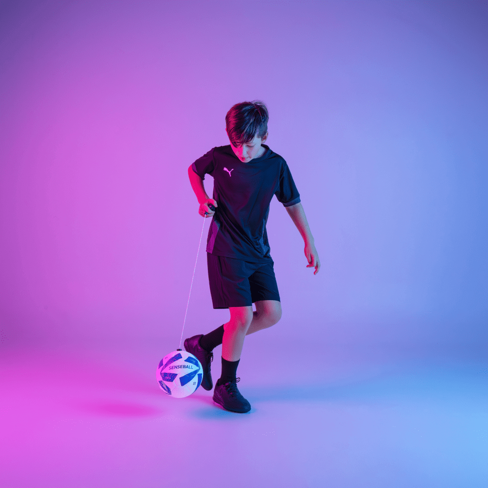 Boy training soccer ball with SenseBall kick trainer on string in blue background